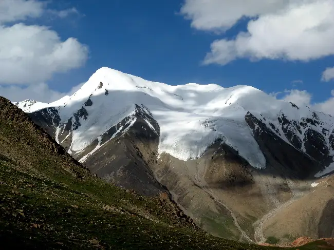 Snow-dusted mountain peak rising sharply against a vibrant blue sky, with a thick glacier and permanent snow cap covering the upper slopes. The foreground shows grassy green alpine meadows and rocky brown lower slopes, creating a striking contrast in the rugged Pamir (or possibly Tian Shan) landscape of Central Asia.