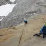 Climber in blue jacket and helmet kneels low on a steep granite slab while alpine rock climbing, with a second roped climber ahead and snowy mountains in the background.