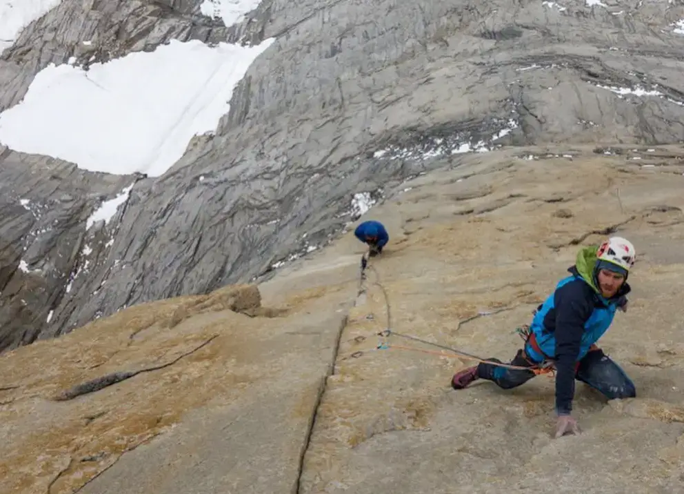 Climber in blue jacket and helmet kneels low on a steep granite slab while alpine rock climbing, with a second roped climber ahead and snowy mountains in the background.