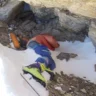 A deceased climber known as "Green Boots" lies curled in a rocky crevice on Mount Everest, wearing a bright orange jacket, blue pants, and distinctive lime-green climbing boots, with an ice axe and oxygen bottles nearby in the snow.