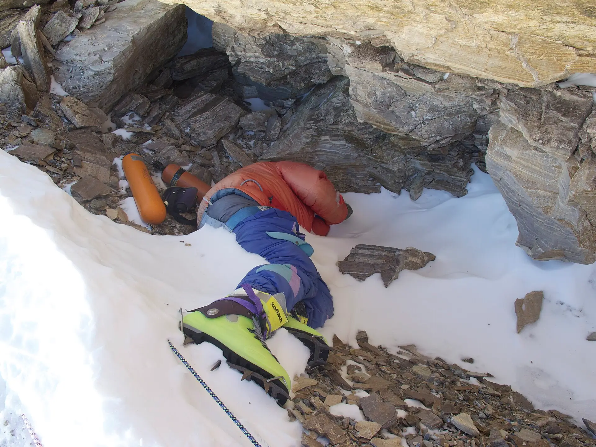 A deceased climber known as "Green Boots" lies curled in a rocky crevice on Mount Everest, wearing a bright orange jacket, blue pants, and distinctive lime-green climbing boots, with an ice axe and oxygen bottles nearby in the snow.