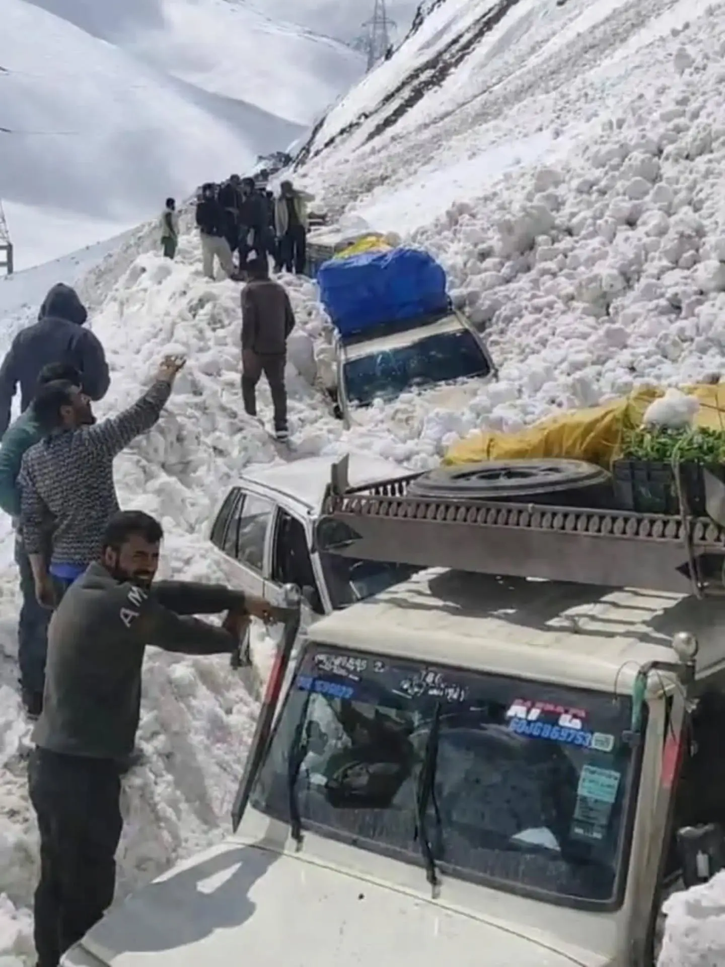 "A group of men standing on a steep, snow-covered mountain slope after a vehicle accident. Several white cars and vans are partially buried or stuck in deep snow, with one vehicle tilted sideways and another loaded with a blue tarp and cargo. People are gathered around the scene, with one man in the foreground gesturing and appearing to direct efforts while others observe from higher up the snowy hill."