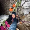 Climber in winter gear taking a selfie inside a snowy rock niche on a big wall route. He's wearing a black balaclava, helmet hanging nearby, surrounded by colorful gear and ropes, with a grin despite the cold, dark conditions.