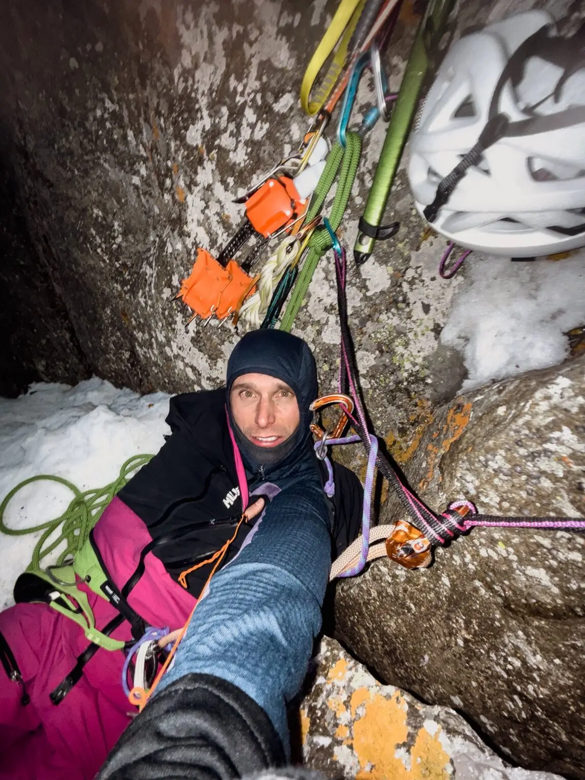 Climber in winter gear taking a selfie inside a snowy rock niche on a big wall route. He's wearing a black balaclava, helmet hanging nearby, surrounded by colorful gear and ropes, with a grin despite the cold, dark conditions.