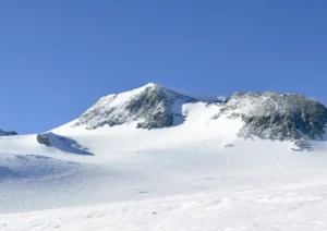 Snow-covered mountain peak under a clear blue sky, with rocky ridges and gentle snowy slopes in the foreground.