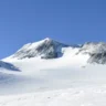 Snow-covered mountain peak under a clear blue sky, with rocky ridges and gentle snowy slopes in the foreground.