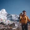 Simone Moro, an Italian mountaineer, stands smiling in the foreground on a rocky Himalayan trail, holding trekking poles. He wears a bright orange jacket and a black cap, with a large backpack. Behind him, the majestic snow-covered peak of Ama Dablam rises sharply against a clear blue sky, with colorful Tibetan prayer flags fluttering in the foreground