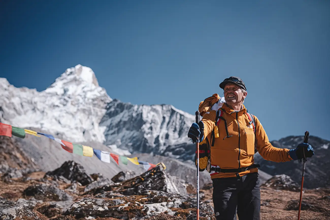 Simone Moro, an Italian mountaineer, stands smiling in the foreground on a rocky Himalayan trail, holding trekking poles. He wears a bright orange jacket and a black cap, with a large backpack. Behind him, the majestic snow-covered peak of Ama Dablam rises sharply against a clear blue sky, with colorful Tibetan prayer flags fluttering in the foreground
