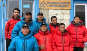 A group of 11 Nepali men, mostly Sherpas, posing together and smiling in front of the blue wooden building of the Sagarmatha Pollution Control Committee (SPCC) office. They wear colorful puffy down jackets—several in bright red, others in blue—with black caps featuring a logo. The front row has four men kneeling or crouching, while the back row of seven stands; one man in the front gives a thumbs-up gesture. A wooden sign above the open door reads "SAGARMATHA POLLUTION CONTROL COMMITTEE" and "SPCC" with "Open" posted below. The scene appears to be at high altitude in the Everest region, likely near Lukla or a related base.