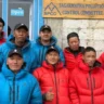 A group of 11 Nepali men, mostly Sherpas, posing together and smiling in front of the blue wooden building of the Sagarmatha Pollution Control Committee (SPCC) office. They wear colorful puffy down jackets—several in bright red, others in blue—with black caps featuring a logo. The front row has four men kneeling or crouching, while the back row of seven stands; one man in the front gives a thumbs-up gesture. A wooden sign above the open door reads "SAGARMATHA POLLUTION CONTROL COMMITTEE" and "SPCC" with "Open" posted below. The scene appears to be at high altitude in the Everest region, likely near Lukla or a related base.