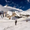 Three mountaineers trek across the snowy Khumbu Icefall on Mount Everest's route in Nepal. Two climbers in the foreground carry a long aluminum ladder horizontally on their shoulders, while a third ahead wears a large backpack. They move carefully over the uneven, snow-covered glacier terrain with towering seracs, massive ice walls, and a sharp, rocky peak rising dramatically in the background under a deep blue sky with scattered clouds.