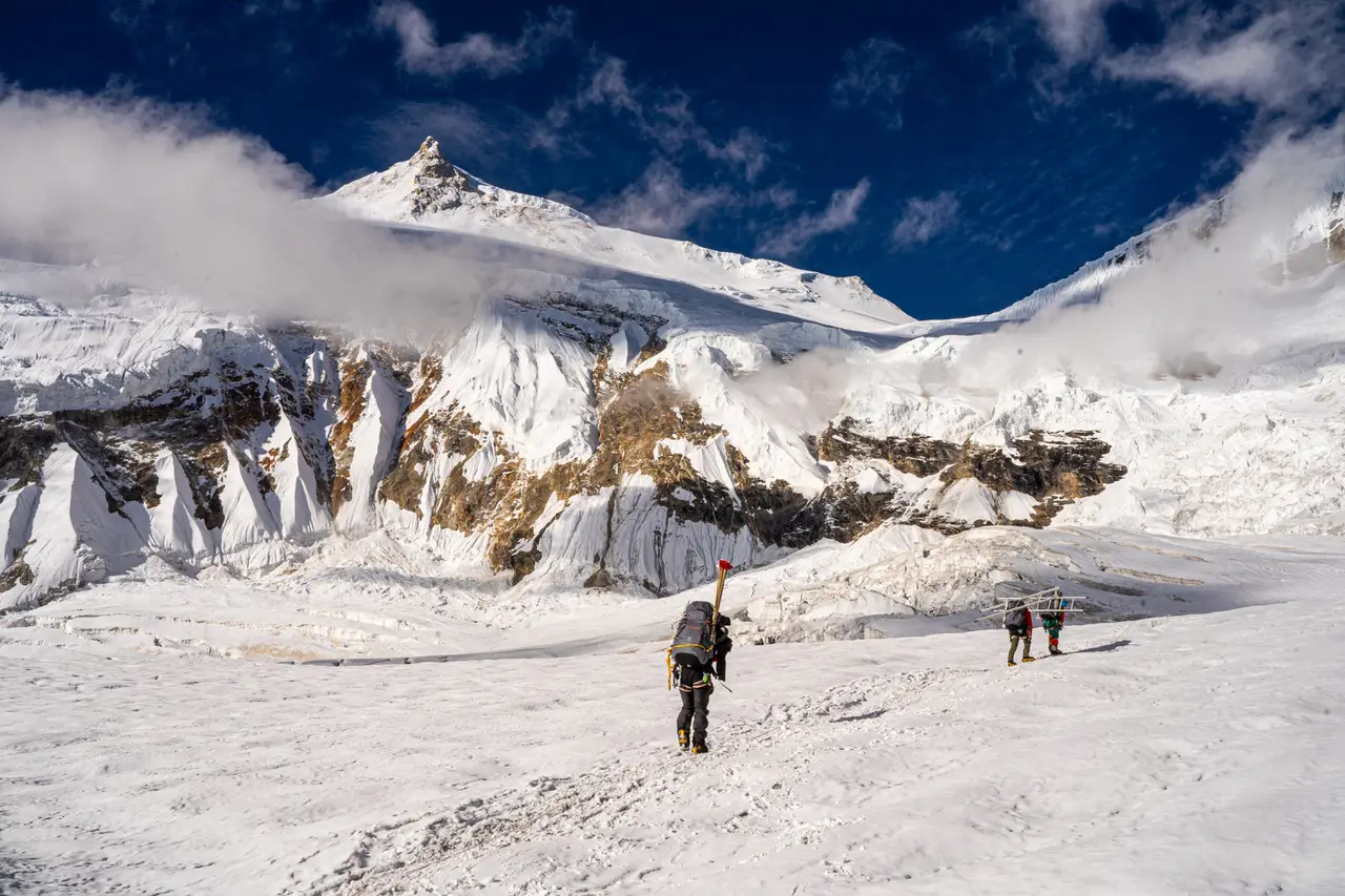 Three mountaineers trek across the snowy Khumbu Icefall on Mount Everest's route in Nepal. Two climbers in the foreground carry a long aluminum ladder horizontally on their shoulders, while a third ahead wears a large backpack. They move carefully over the uneven, snow-covered glacier terrain with towering seracs, massive ice walls, and a sharp, rocky peak rising dramatically in the background under a deep blue sky with scattered clouds.