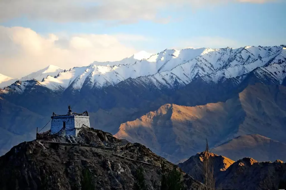 Ancient whitewashed Buddhist stupa or small monastery perched on a rocky hilltop in the Ladakh region, overlooking dramatic snow-capped mountain ranges bathed in warm golden sunset light.