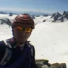 Selfie of a mountaineer in a red-and-black climbing helmet and orange-tinted glacier glasses, smiling at the camera. Behind him stretches a vast snowy glacier and jagged, snow-covered peaks under a clear blue sky.