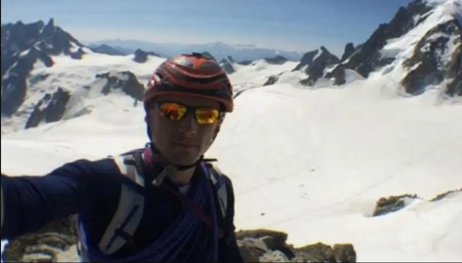 Selfie of a mountaineer in a red-and-black climbing helmet and orange-tinted glacier glasses, smiling at the camera. Behind him stretches a vast snowy glacier and jagged, snow-covered peaks under a clear blue sky.