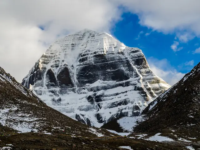 Majestic Mount Kailash (Kangrinboqê) covered in fresh snow, viewed from the front with dark rocky ridges on both sides under a partly cloudy sky.