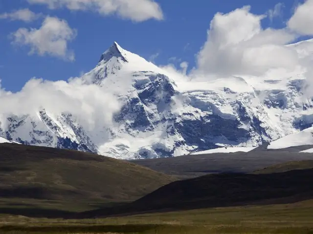 Snow-capped Mount rises sharply against a bright blue sky, surrounded by grassy alpine slopes in the Georgian Caucasus.